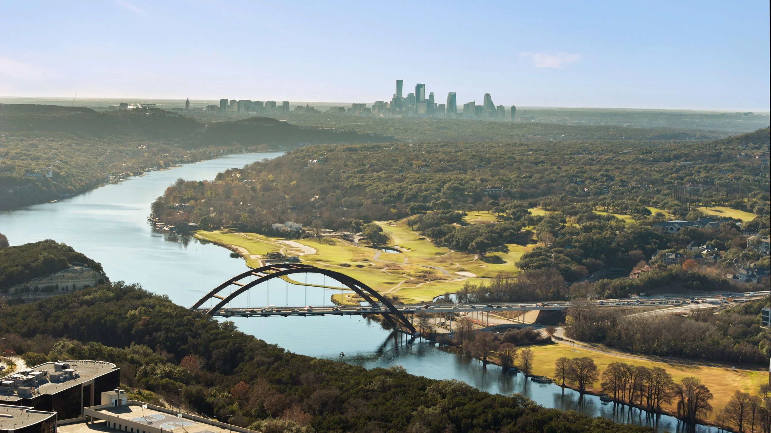 Aerial view of the 360 Bridge over Lake Austin with the Austin skyline beyond, late-afternoon light across the hill country.
